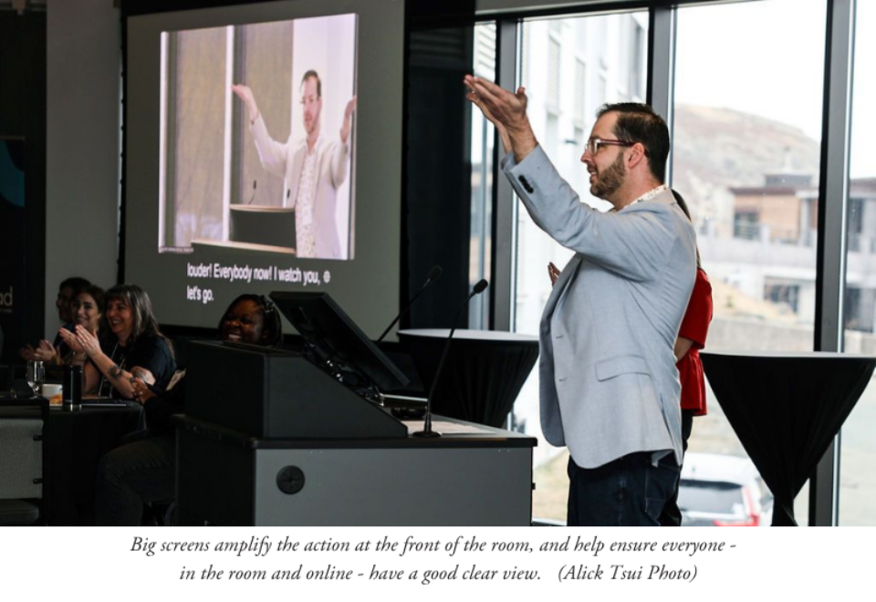 Clean cut bearded person standing at front of room with arms raised with a duplicate image of him on screen to the left of the frame